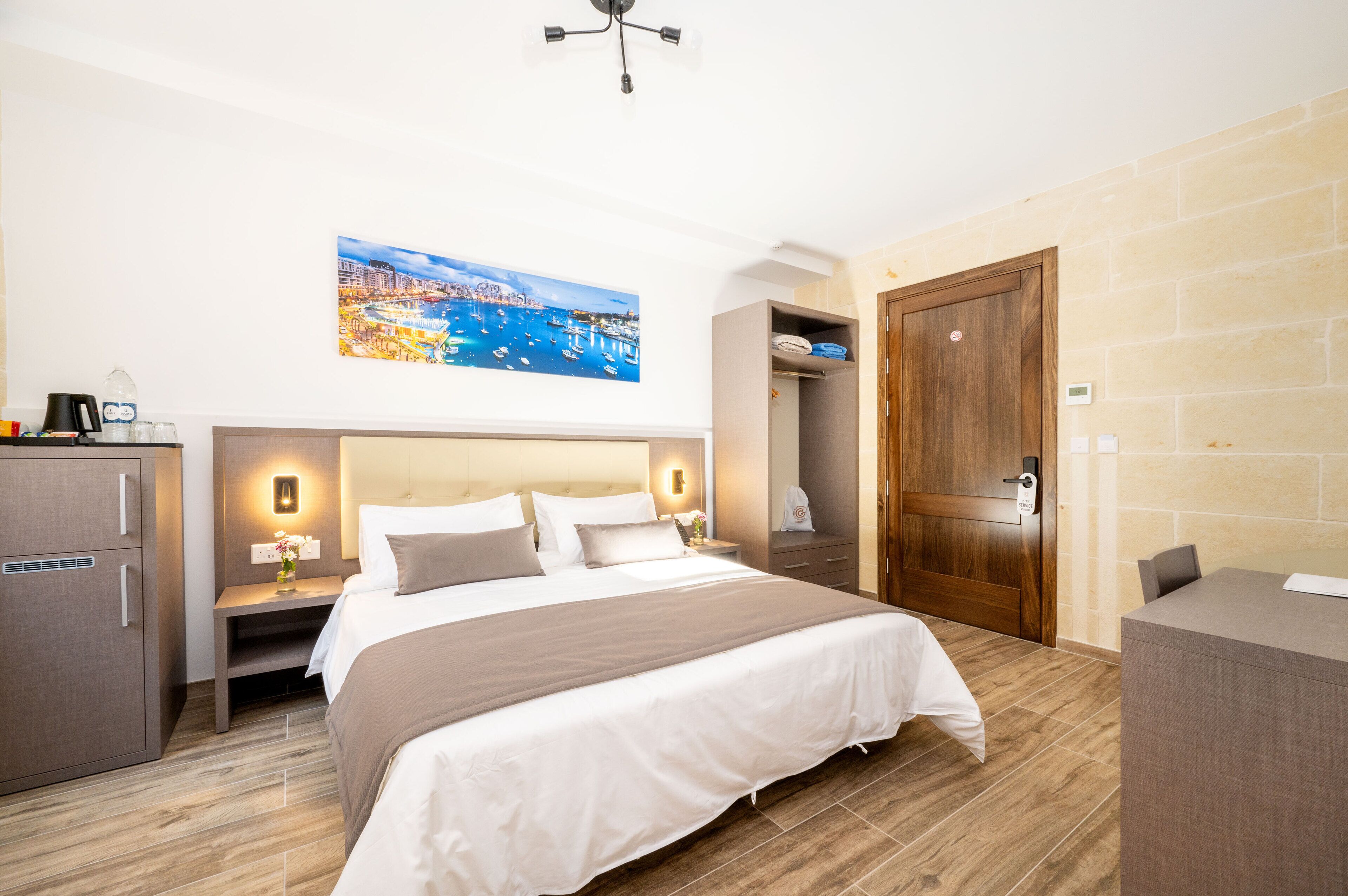 A hotel room with a large bed, a wooden wardrobe, a desk, and a painting of a coastal city.