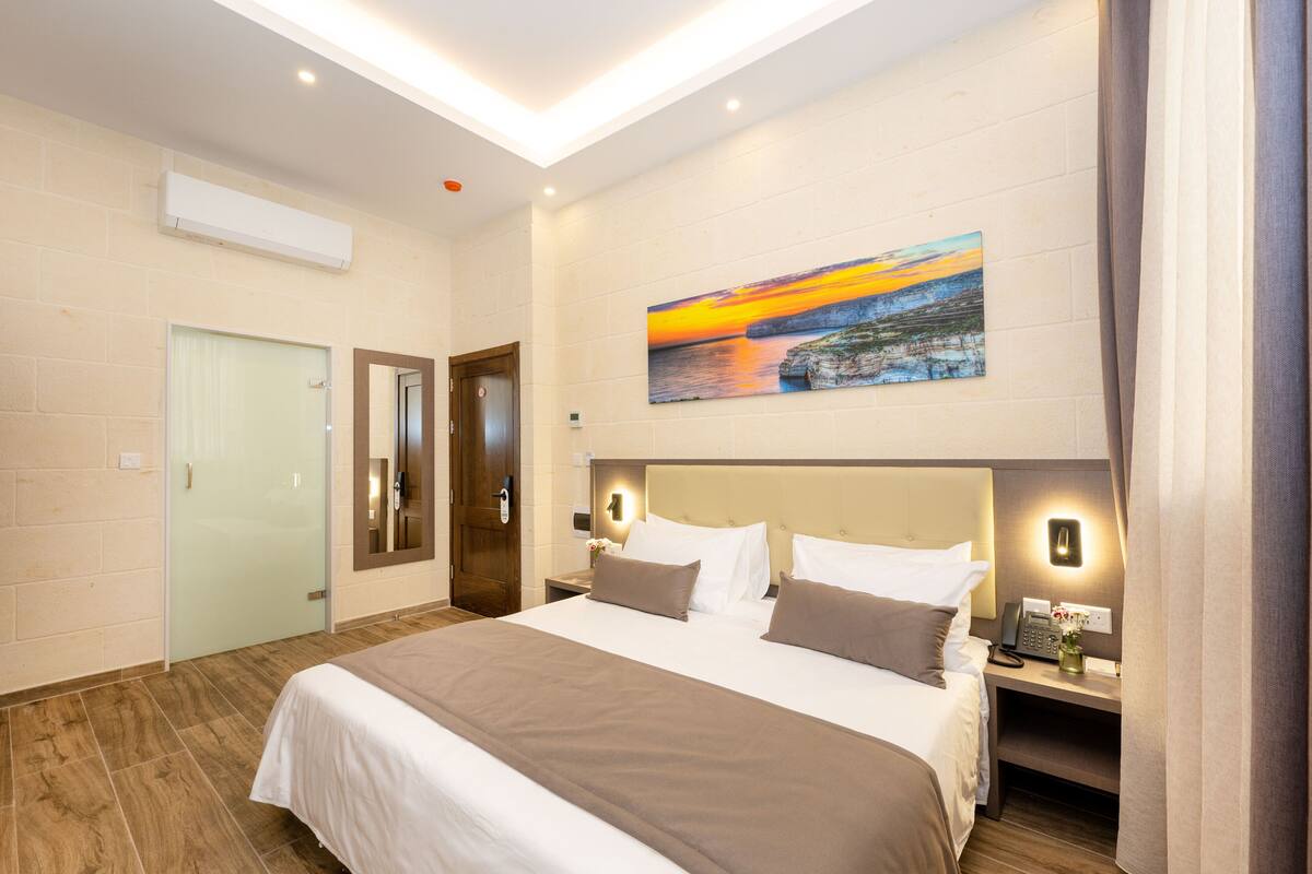 Habitación de hotel con una cama grande, un cuadro de un atardecer, una mesita de noche con un teléfono y un florero, y una puerta con un espejo.