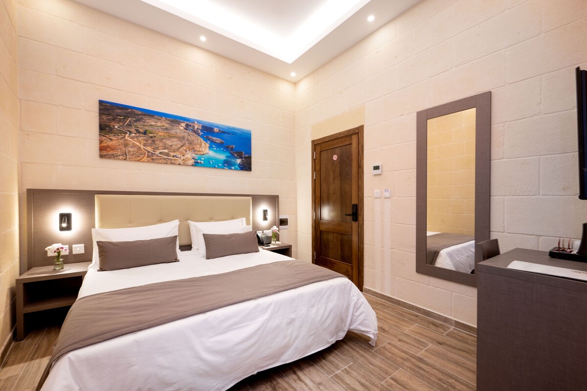 Una habitación de hotel moderna con una cama grande, una mesita de noche con lámpara, una puerta de madera y un espejo amplio.