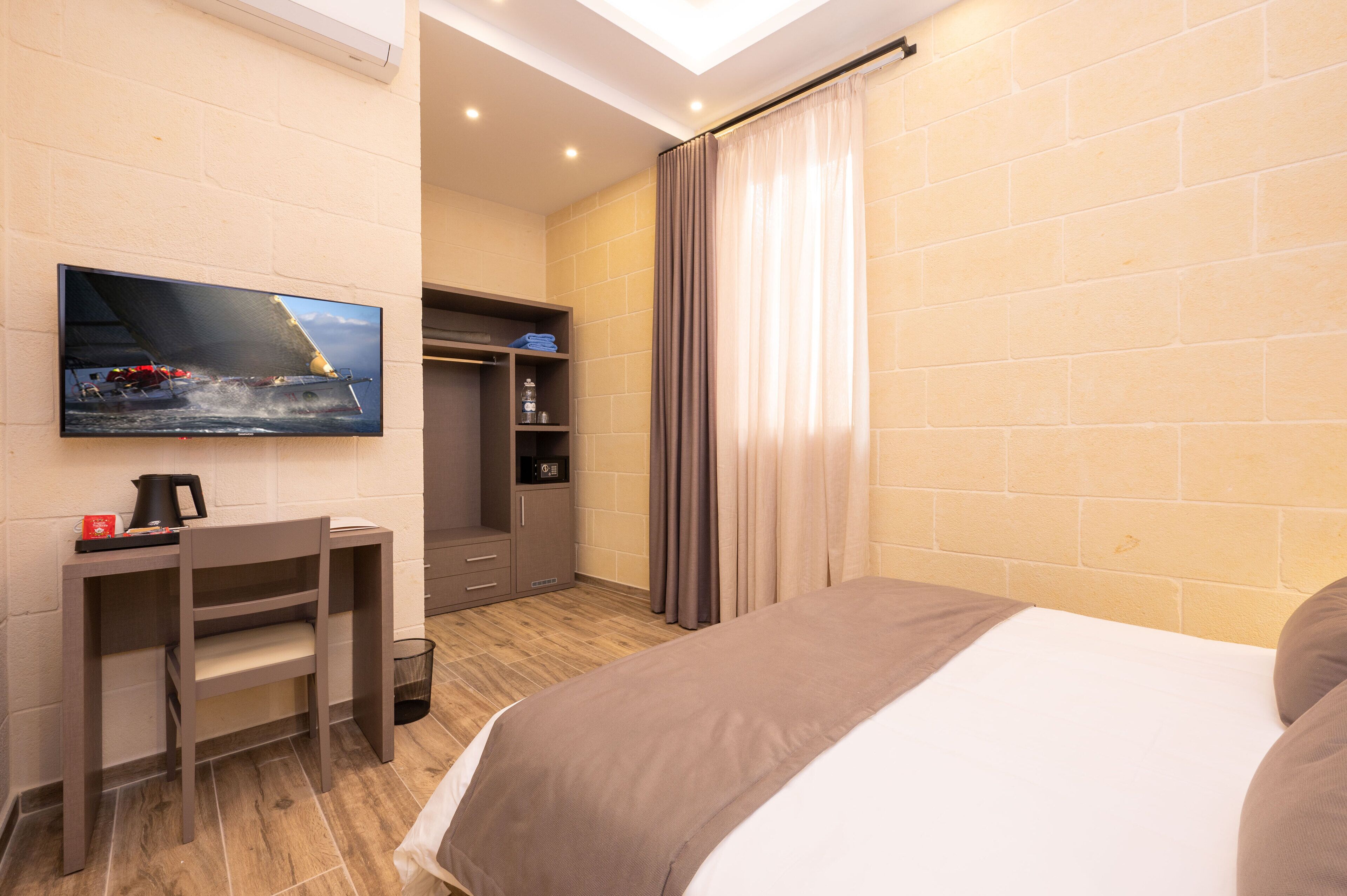A hotel room with a bed, a desk with a chair, a TV mounted on the wall, a wardrobe, and a bathroom visible through an open door.