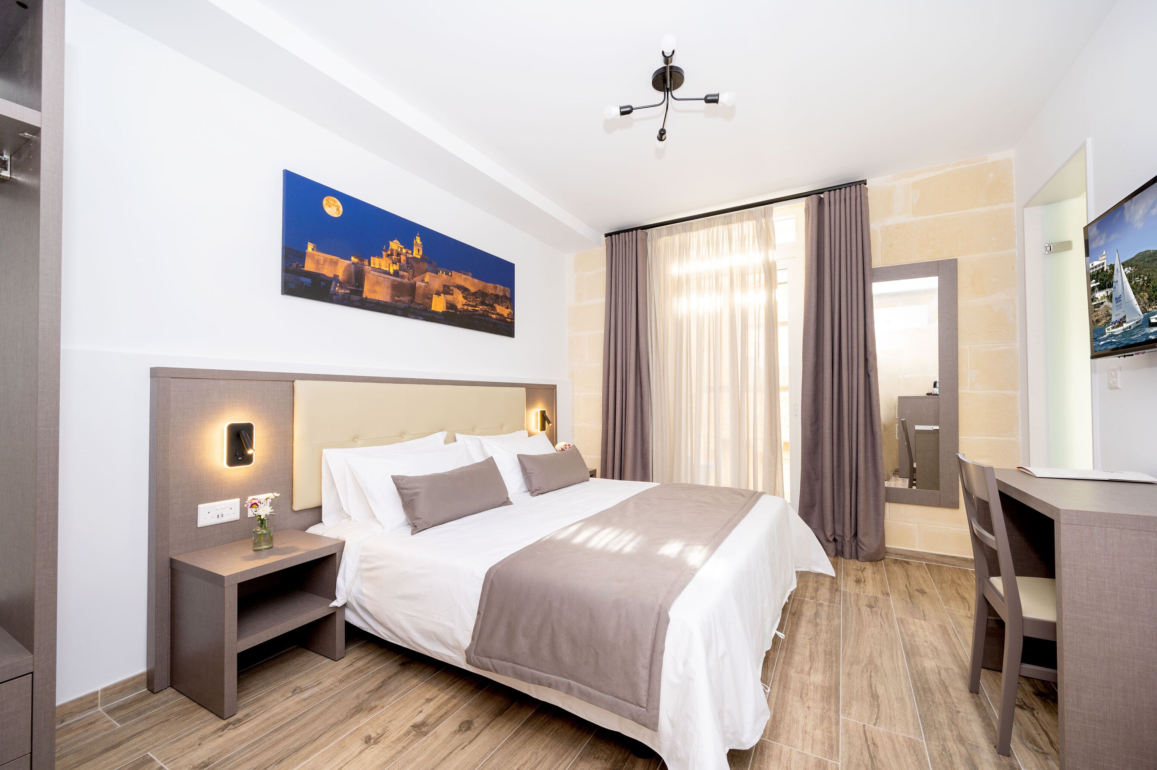 A modern hotel room with a large bed, bedside tables, a desk, a TV, and a painting of a cityscape.
