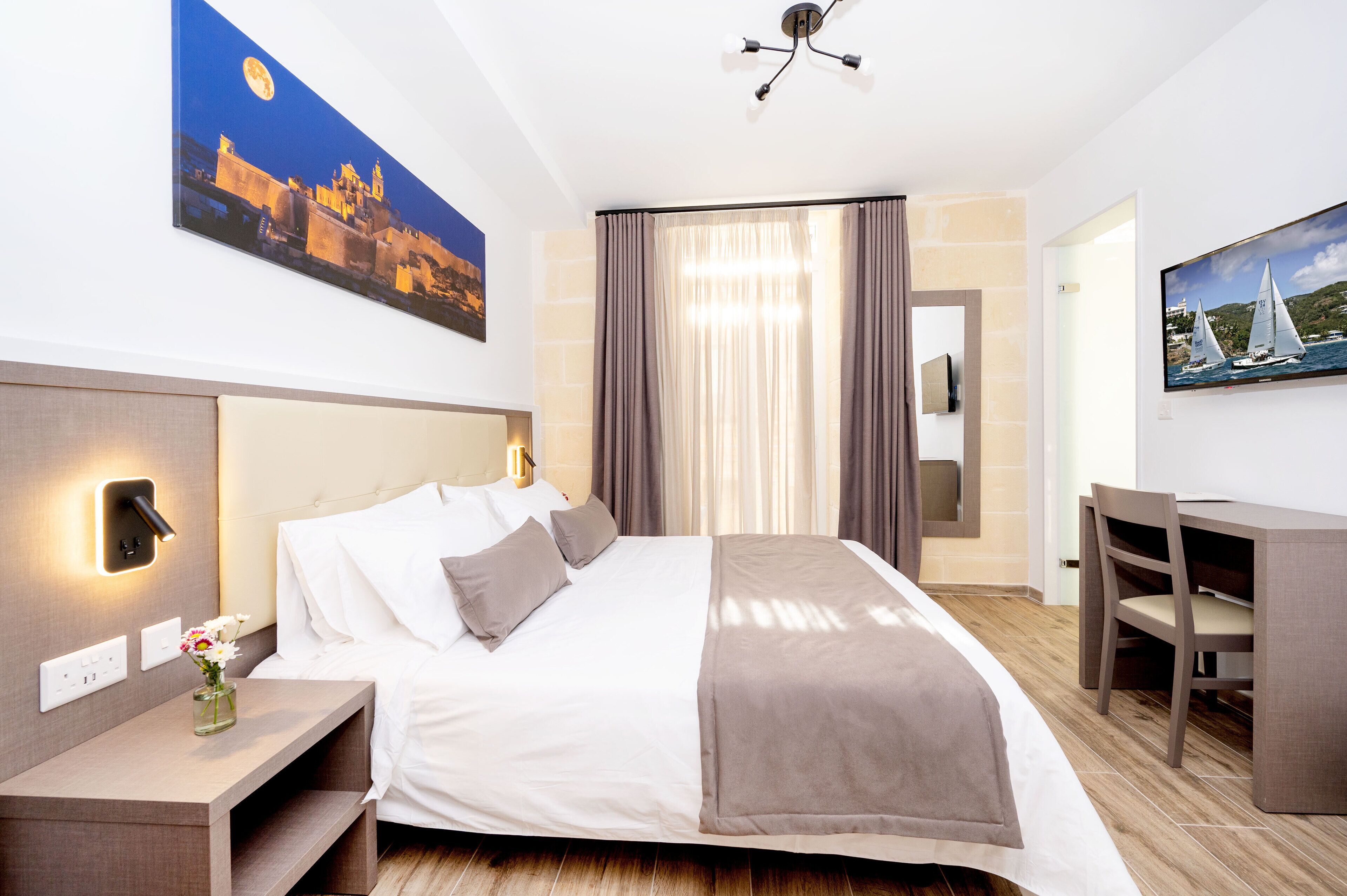 Une chambre d’hôtel avec un lit, une table de chevet et un bureau avec une chaise. Il y a deux tableaux accrochés aux murs et un luminaire au plafond.