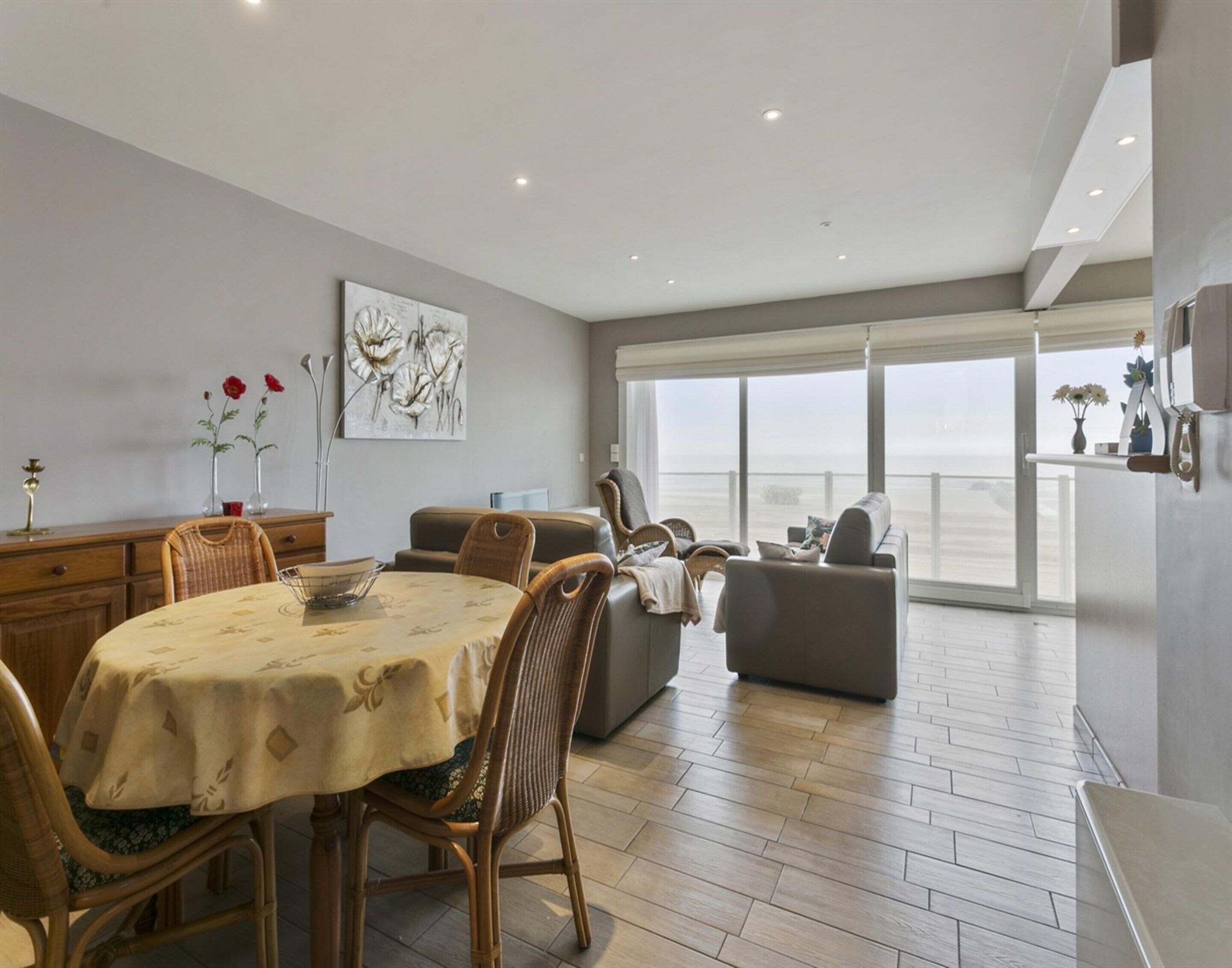 A spacious living area with a dining table, chairs, and a sofa. There is a large window offering a view of the sea.