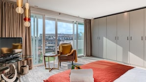 A modern hotel room with a large bed, a TV, a seating area with a chair and table, and a balcony with a view.