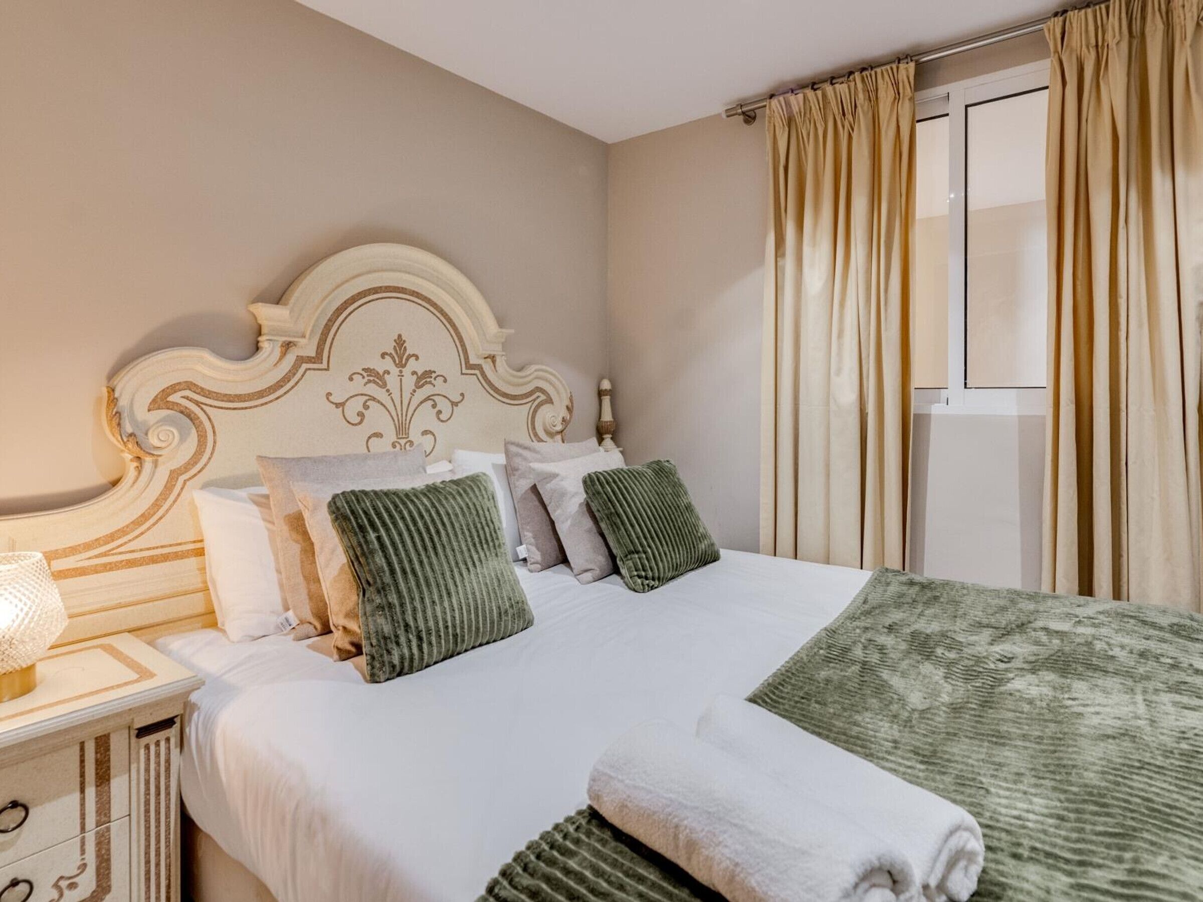A neatly made bed with a white comforter and green pillows, a bedside lamp, and a window with beige curtains.