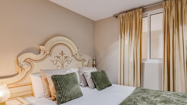 A neatly made bed with a white comforter and green pillows, a bedside lamp, and a window with beige curtains.