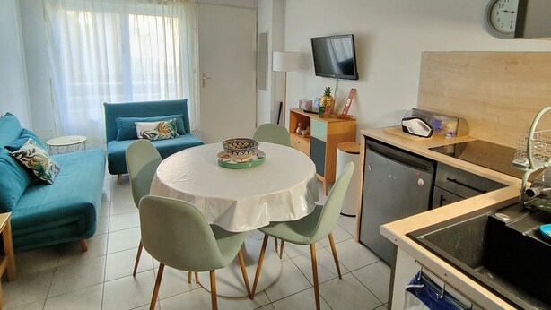 A compact living space with a dining table, chairs, a sofa, a television, a clock, and a kitchenette.