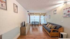 A cozy living room with a sofa, a dining table, and chairs. There are posters on the walls and a ceiling light.