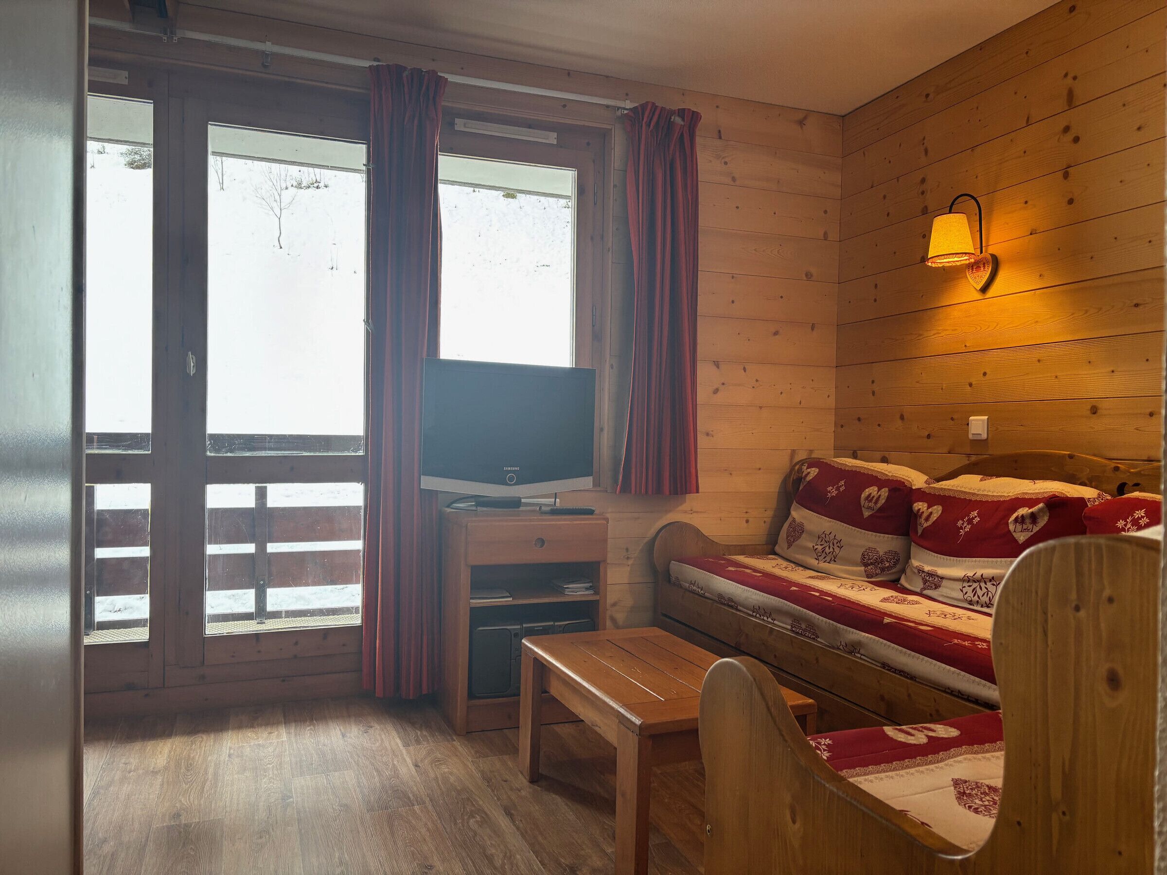 A cozy room with a wooden interior, a sofa with patterned cushions, a television on a wooden stand, and a window with curtains.