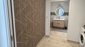 Une salle de bain moderne avec un mur orné d’un motif géométrique, un miroir rond, un lavabo et un lave-linge.