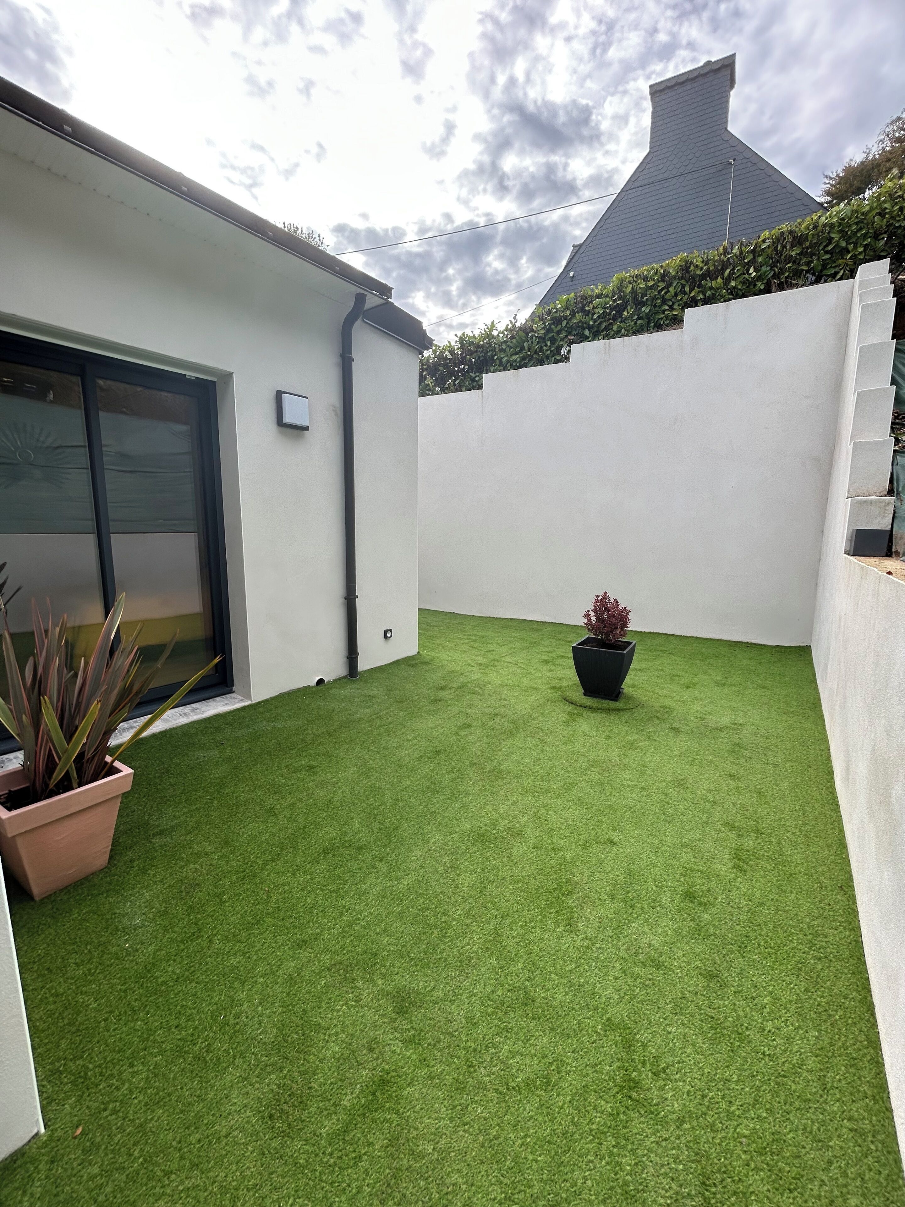 A modern house with a flat roof, a white exterior, and a small backyard with artificial grass and potted plants.