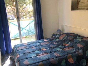 A bedroom with a bed, a window with blue curtains, and a view of a green outdoor area. - Splendid renovated T3 with pool and parking 100m from sea, 6 pers, terrace, WIFI (Cap d'Agde)
