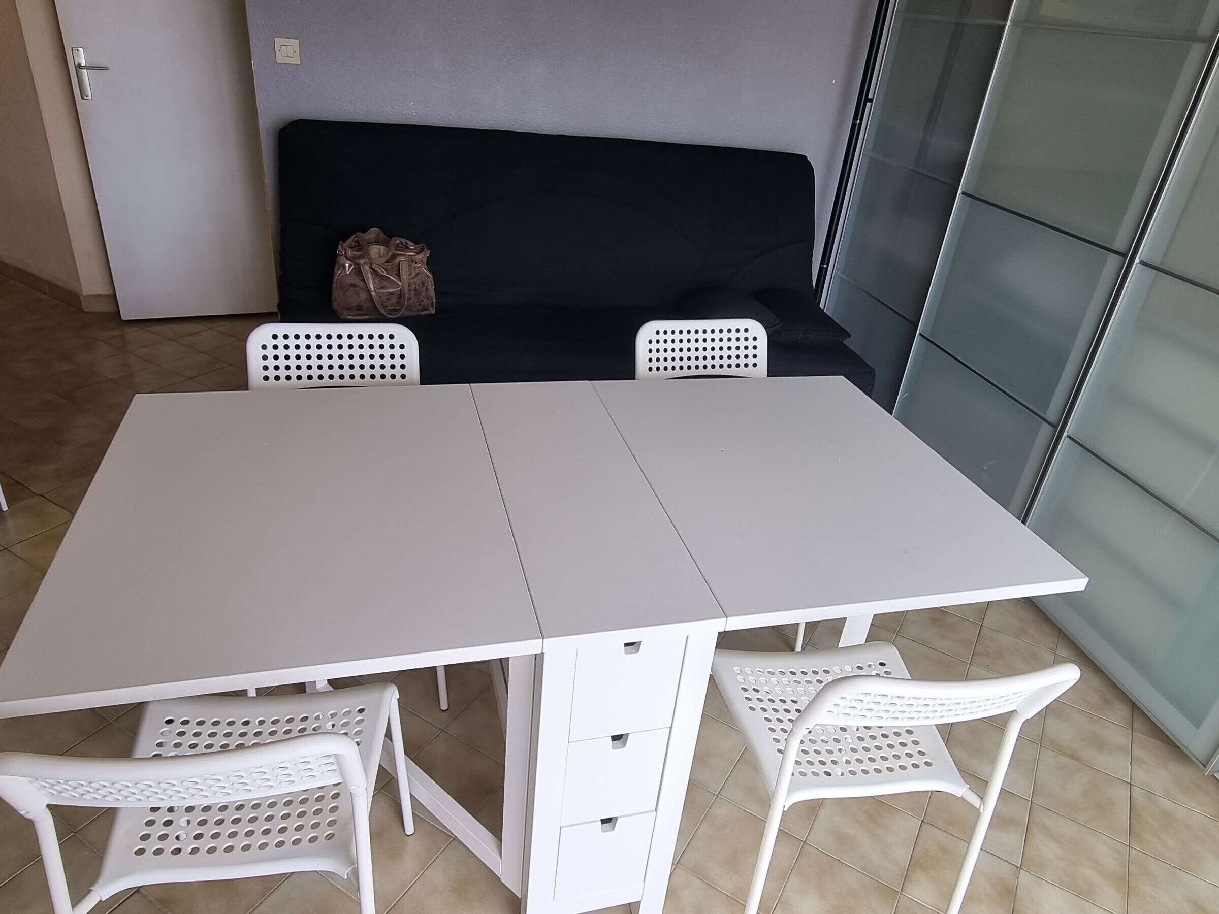 Un'area pranzo con un tavolo bianco e quattro sedie bianche. C'è un divano nero appoggiato al muro e una borsa bianca sopra.