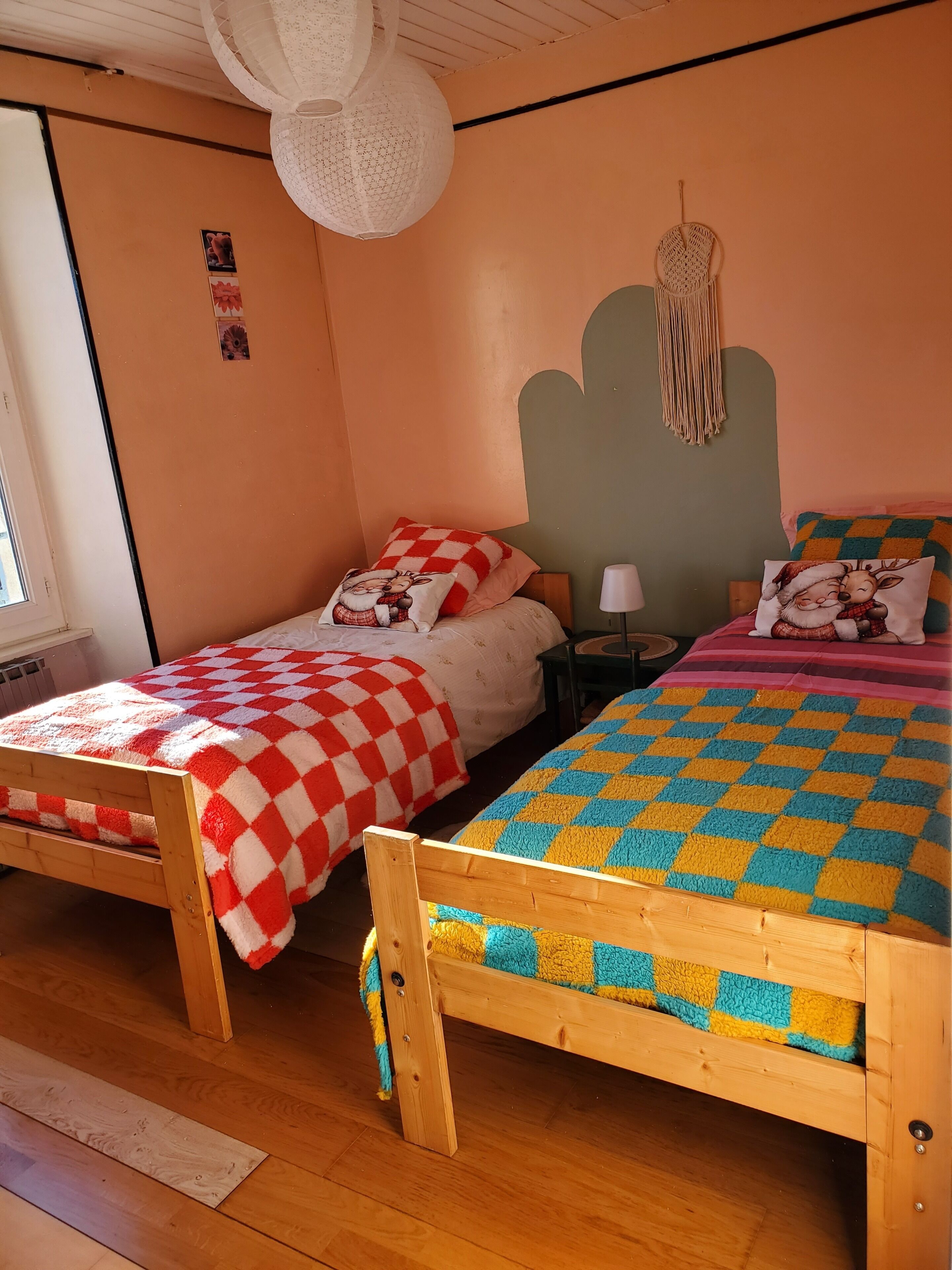 A room with two wooden beds, each with a checkered blanket. There is a white hanging light fixture and a macrame wall hanging.