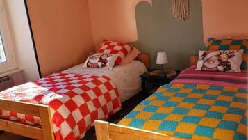 A room with two wooden beds, each with a checkered blanket. There is a white hanging light fixture and a macrame wall hanging.