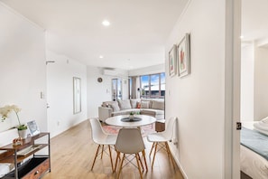 Uma sala de estar moderna com sofá, mesa de jantar e cadeiras, e cozinha visível por uma porta aberta.