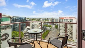A balcony with a glass table and chairs, overlooking a cityscape with modern buildings and a clear sky.