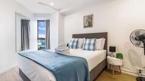 A hotel room with a bed, a bedside table with a lamp, a fan, and a view of the outside.