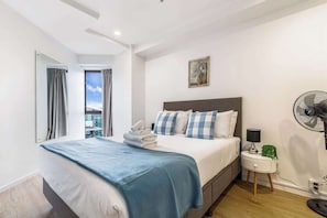 Hotellihuone, jossa on sänky, yöpöytä lampun kanssa, tuuletin ja näkymä ulos.