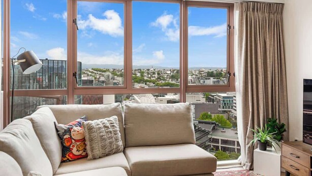 A modern living room with a large window offering a city view, a sofa with decorative pillows, a floor lamp, and a small table with a plant.