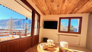 Een gezellige kamer met houten lambrisering, een groot raam met uitzicht op een besneeuwd landschap, een houten tafel met boeken en een glas, en een tv aan de muur.