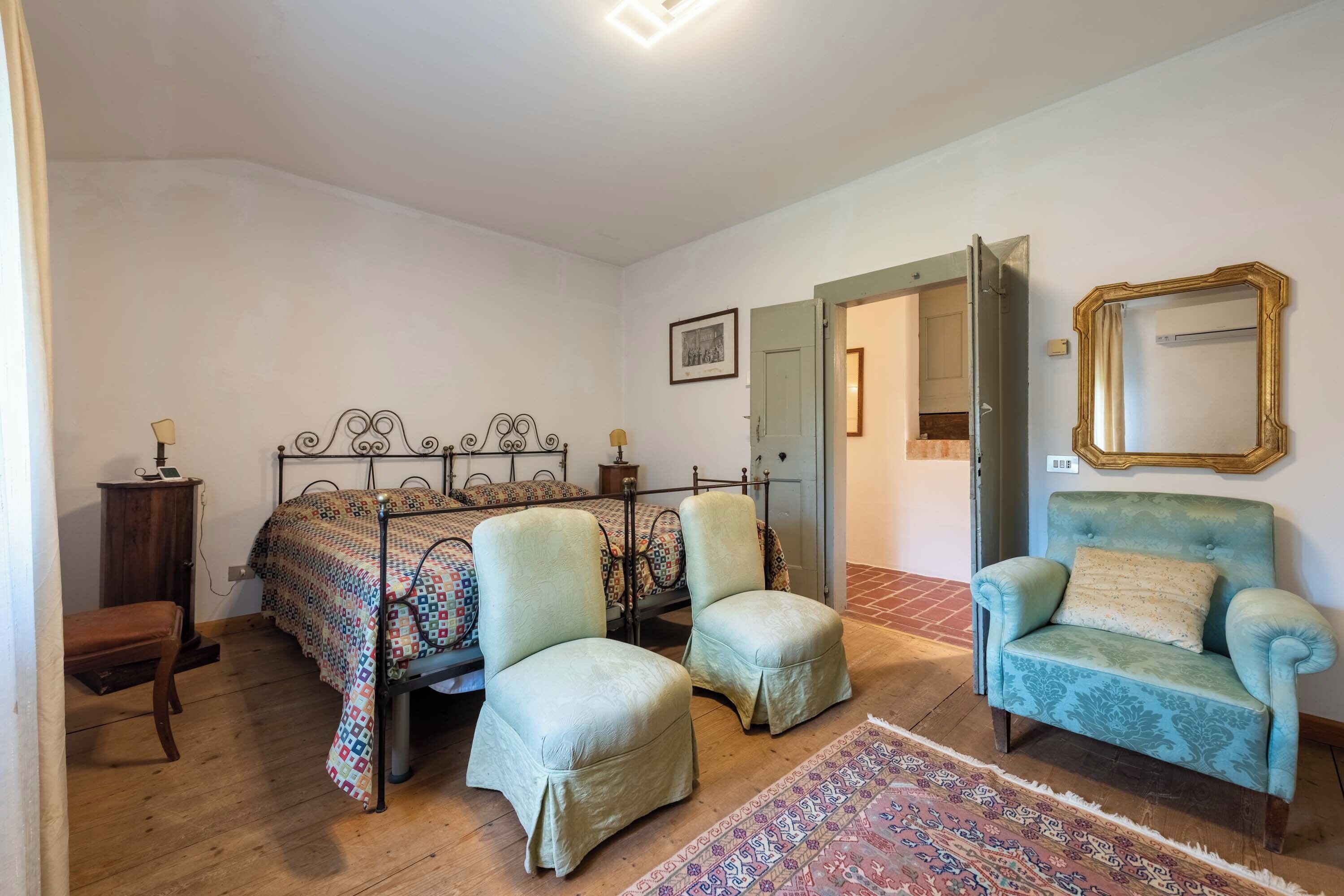 Un dormitorio con una cama, dos sillones, un banco, una alfombra, un espejo y una puerta que da a otra habitación.