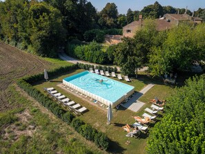 An aerial view of a rectangular swimming pool surrounded by lounge chairs and umbrellas, with a pathway leading to a building in the background.