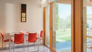 Un comedor con mesa de vidrio y sillas rojas, un cuadro en la pared y vista a un jardín a través de una puerta corrediza de vidrio.