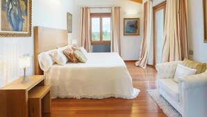 Un dormitorio con cama, cabecera de madera, mesita de noche, sofá blanco y un ventanal con cortinas.