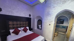 A bedroom with a large bed, a headboard with a decorative pattern, a red and white bedspread, and a small window with a view. - blue city (Chefchaouen)