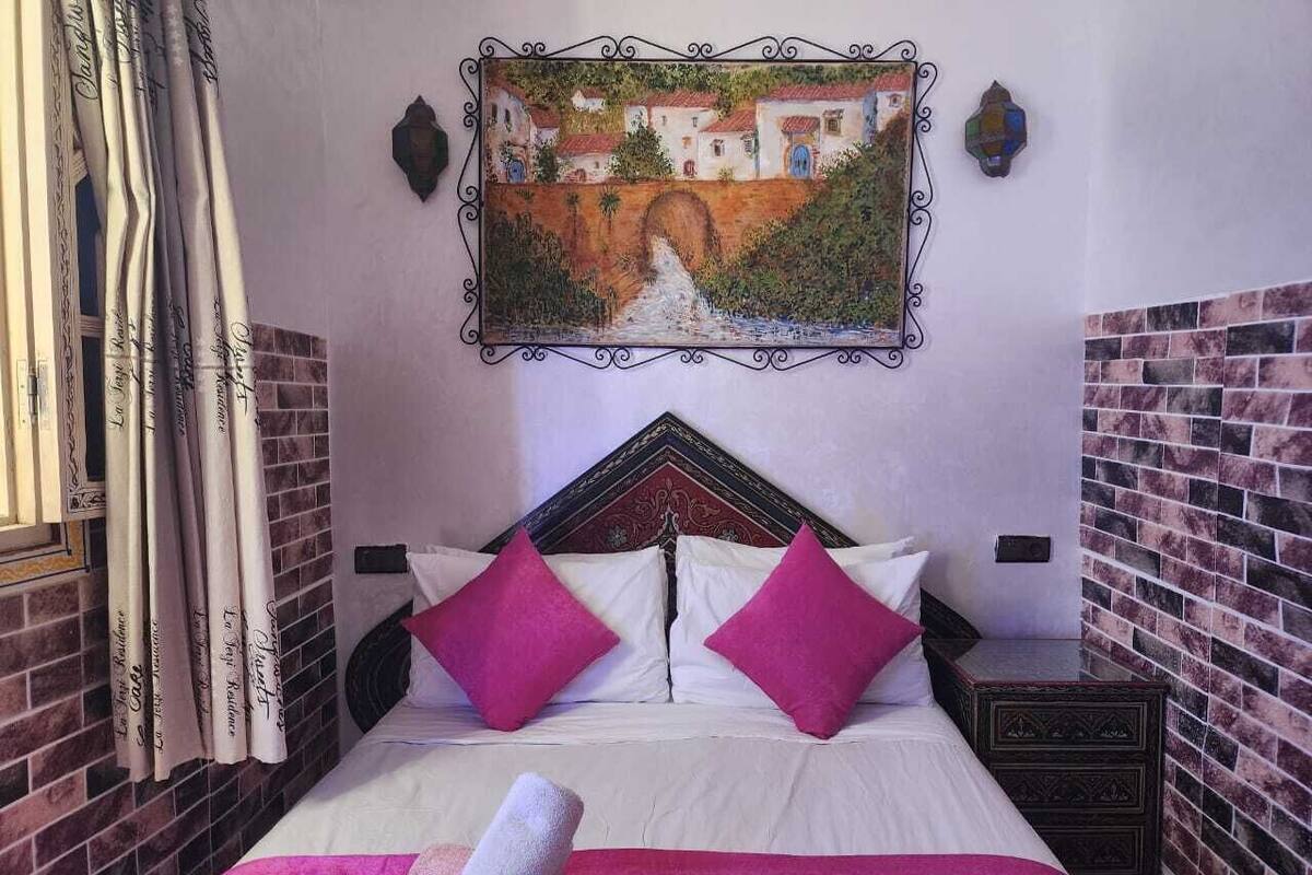 A hotel room with a bed, two pillows, a nightstand, a painting, and a window with curtains.