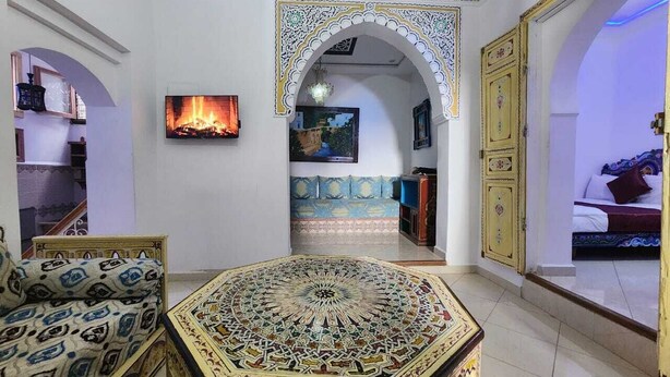 A room with a large, ornate rug, a sofa, a television, and a bed with a blue and white patterned bedspread. - blue city (Chefchaouen)