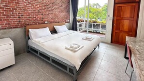 Une chambre d’hôtel avec un mur en briques, un lit recouvert de draps blancs, une porte en bois et un balcon donnant sur un paysage verdoyant.