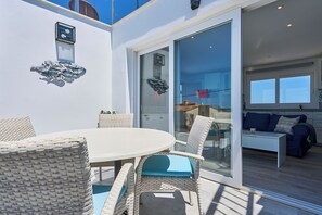 A modern outdoor dining area with wicker furniture, a glass table, and a sliding glass door leading to a living room with a sofa and a coffee table.