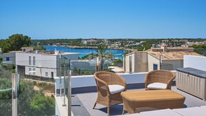Un balcón con muebles de mimbre que da a un pueblo costero y un mar azul cristalino.
