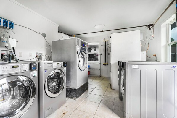 Une buanderie équipée de plusieurs machines à laver et de plusieurs sèche-linge, avec un sol carrelé et une fenêtre donnant sur un paysage agréable.
