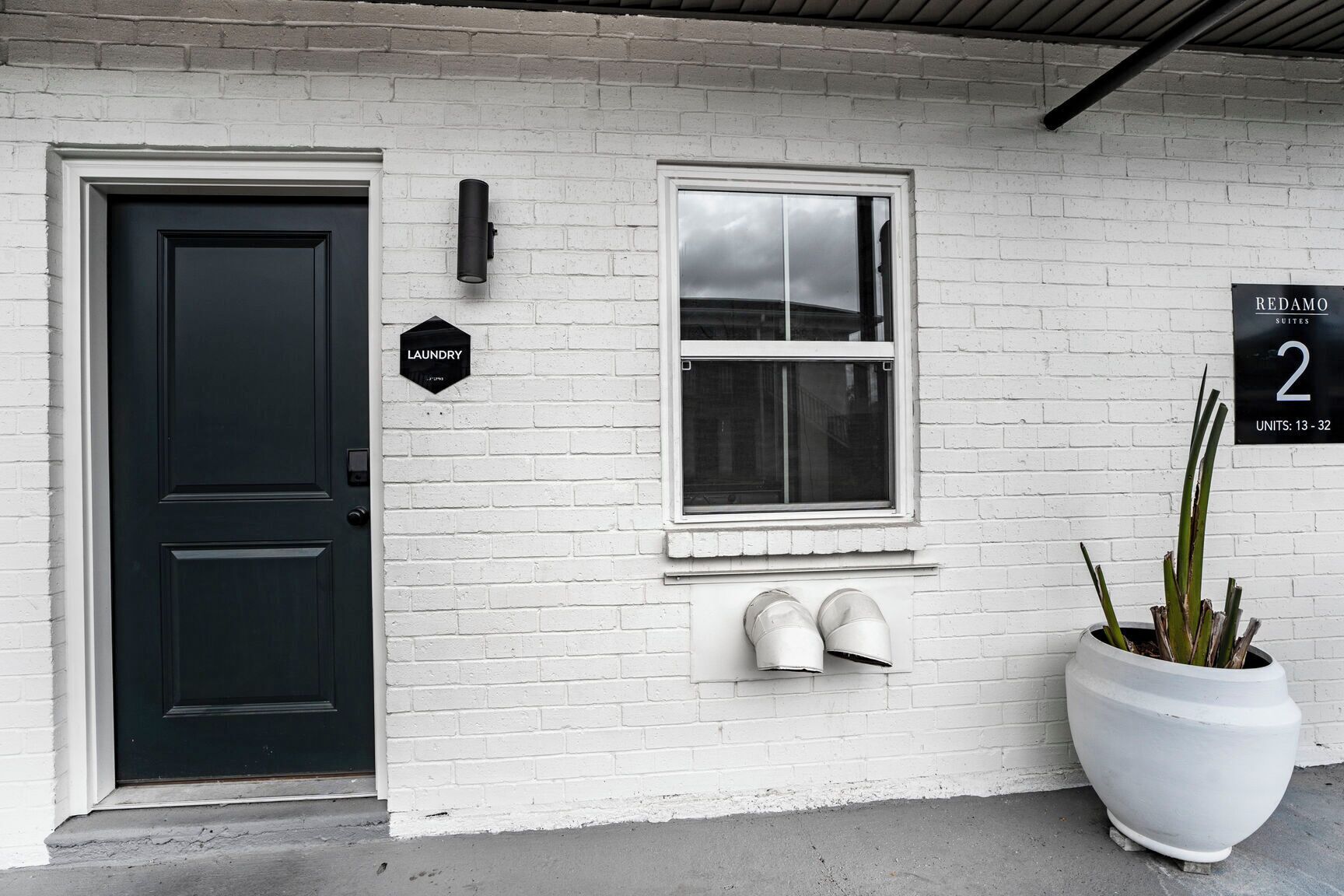 A white brick building with a black door, a window, and a sign indicating "Laundry" and "Redamo Suites 2 Units: 13-32".