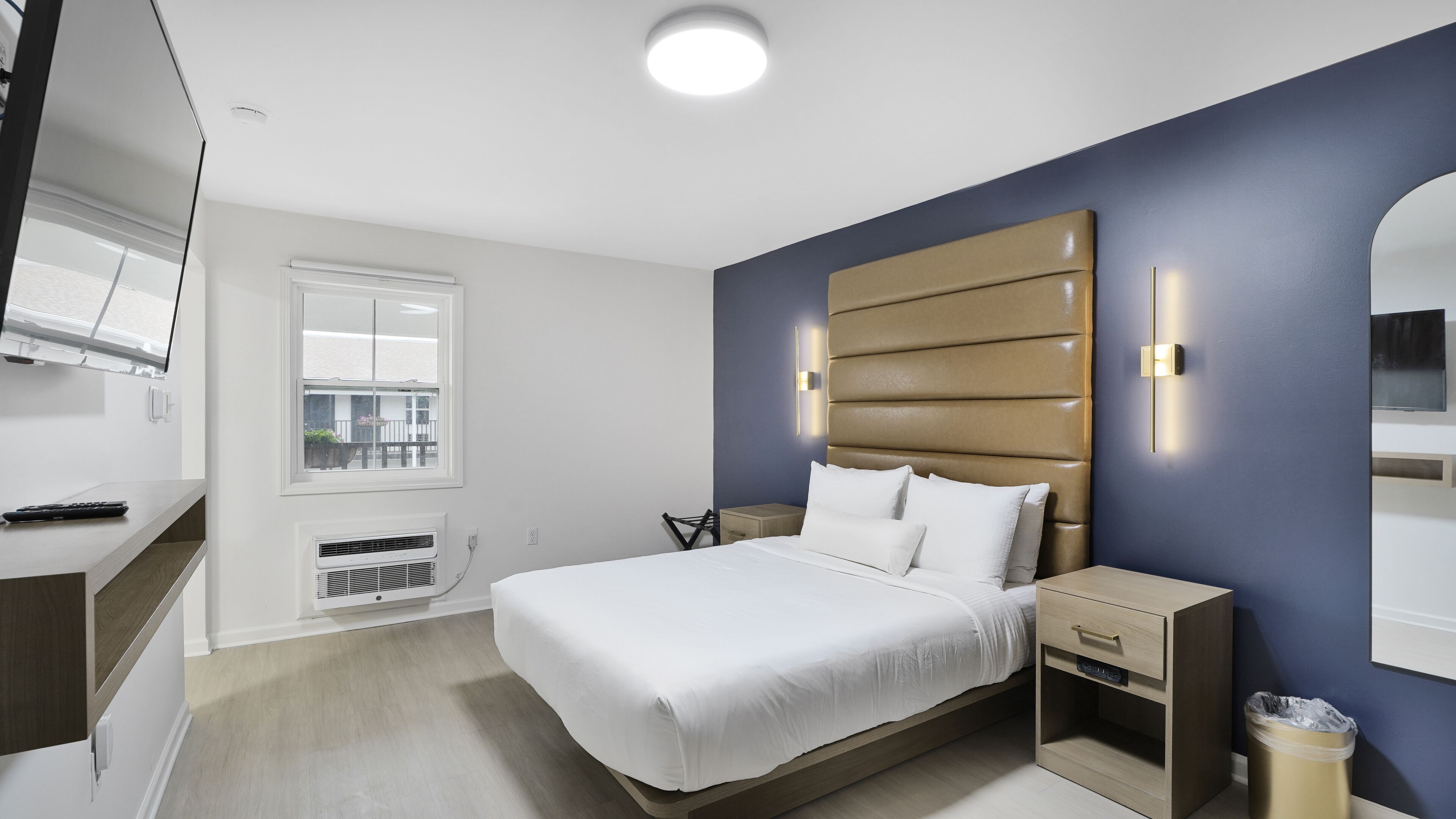 A modern hotel room with a large bed, a bedside table, a nightstand, a wall-mounted TV, and a window with a view of the outside.