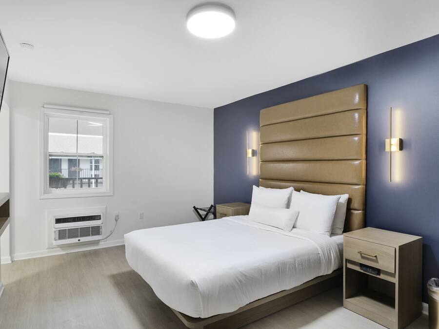 A modern hotel room with a large bed, a bedside table, a nightstand, a wall-mounted TV, and a window with a view of the outside.