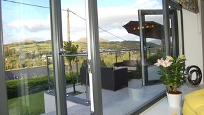 En balkon med glasdør, en potteplante og udsigt over et grønt landskab med bakker.