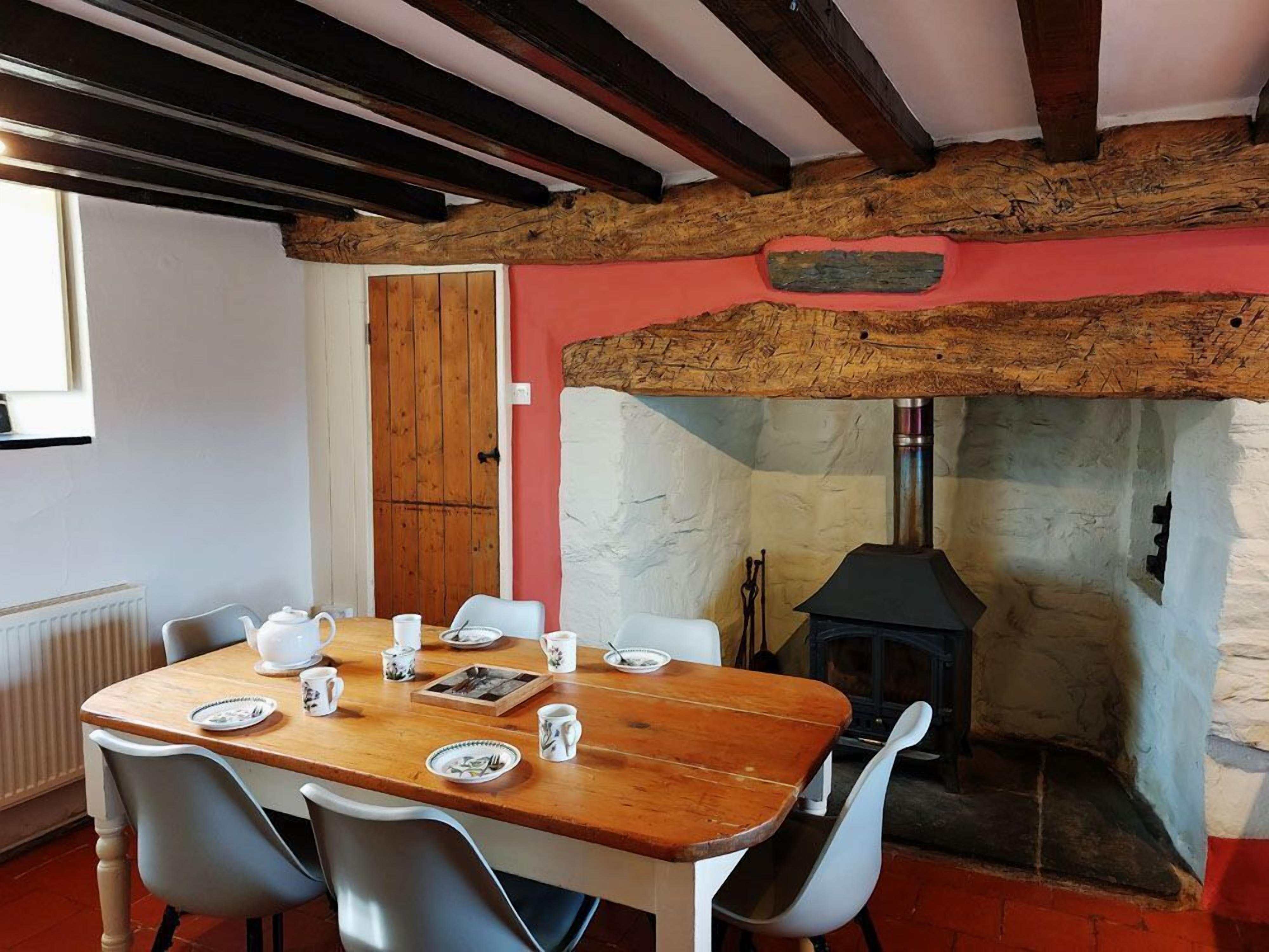 Un comedor con una mesa de madera dispuesta para cuatro personas, rodeada de sillas blancas. Se observa una chimenea de piedra con estufa negra al fondo.