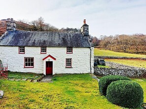 Una casa blanca con puerta roja y varias ventanas, rodeada de un muro de piedra y un patio cubierto de césped.