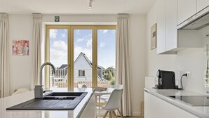 Eine moderne Küche mit einer zentralen Kochinsel, einem Essbereich mit weißem Tisch und Stühlen sowie großen Fenstern mit Blick auf ein Haus und den Himmel.