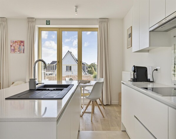 Una cocina moderna con una isla central, un comedor con mesa y sillas blancas, y ventanas amplias que ofrecen vistas a una casa y al cielo.