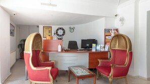 Un área de recepción con un mostrador, dos sillones tapizados en rojo, una mesita y un reloj de pared.