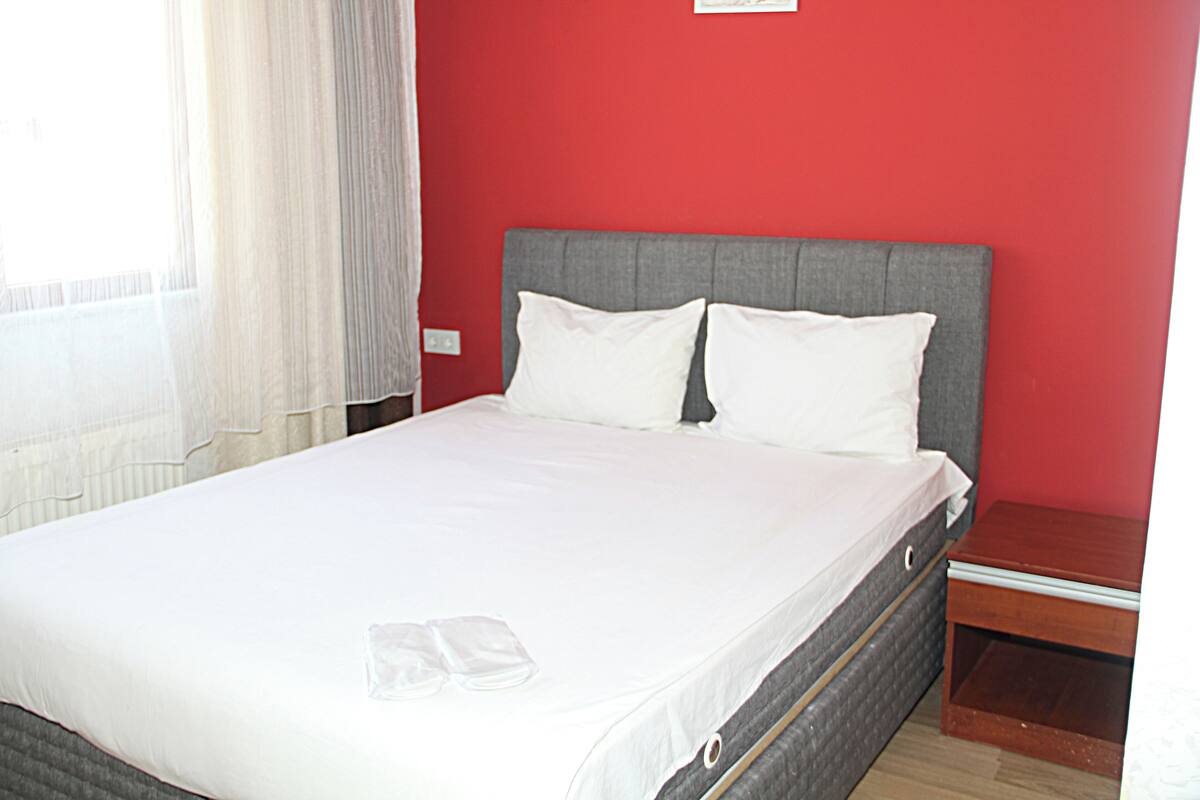 A neatly made bed with white linens and two pillows, a wooden nightstand with a drawer, and a red wall.