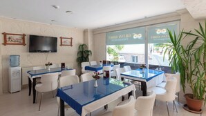Un comedor de hotel con mesas de color azul, sillas blancas, un televisor de pantalla plana y amplias ventanas con vistas a la calle.