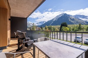 Un balcon doté d’un plancher en bois, de chaises en métal, d’une table et offrant une vue sur des montagnes enneigées.