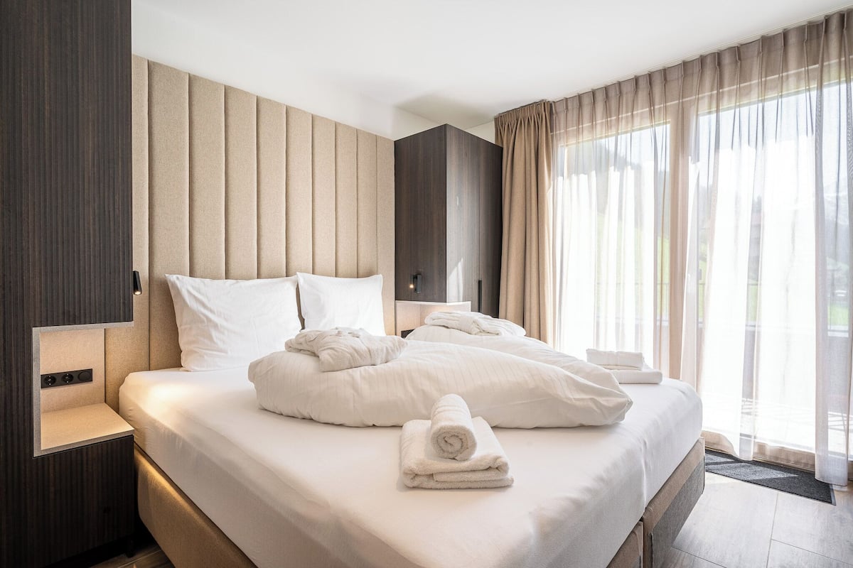 A modern hotel room with a large bed, a bedside table, and a view of the outdoors through sheer curtains.