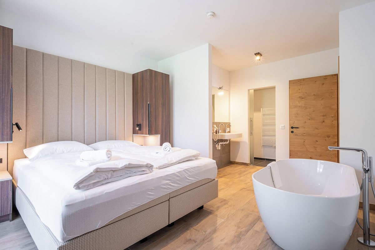 A modern bedroom with a large bed, a white bathtub, and wooden paneling on the walls.